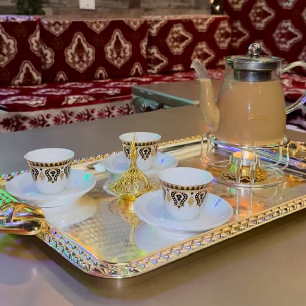 Middle-eastern tea set with a teapot and three ornate cups on a silver tray, with patterned seating in the background.