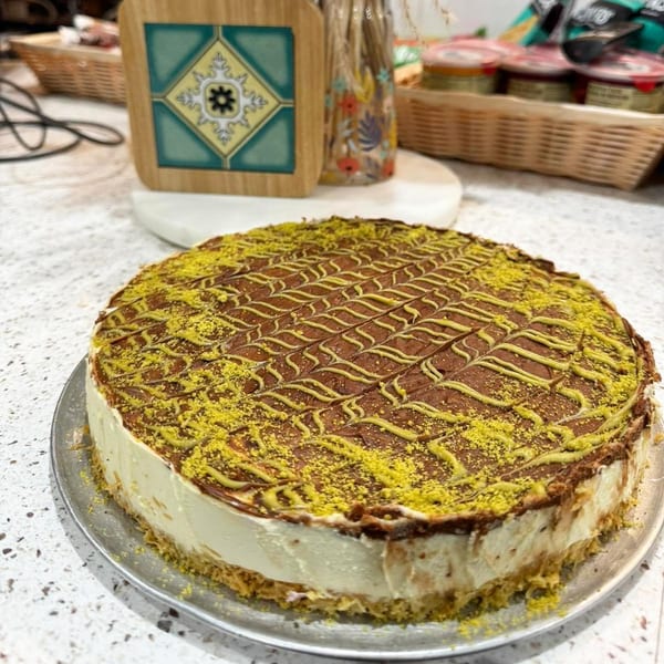 A whole cheesecake with a chocolate top, pistachio powder, and yellow icing swirls on a metal tray.